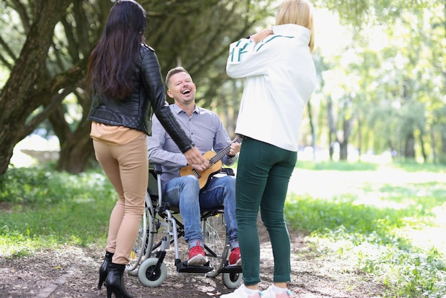 Homem falecido feliz em cadeira de rodas tocando ukulele para seus amigos no parque duas mulheres jovens