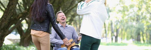 Homem falecido feliz em cadeira de rodas tocando ukulele para seus amigos no parque duas mulheres jovens