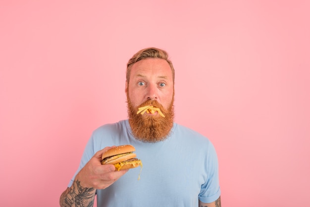 Homem faminto com barba e tatuagens comendo um sandwitch com hambúrguer e batatas