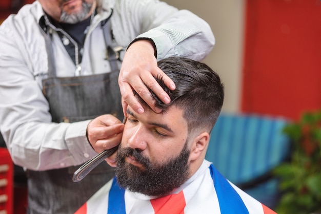 Homem farpado considerável que obtém o corte de cabelo pelo cabeleireiro na barbearia.