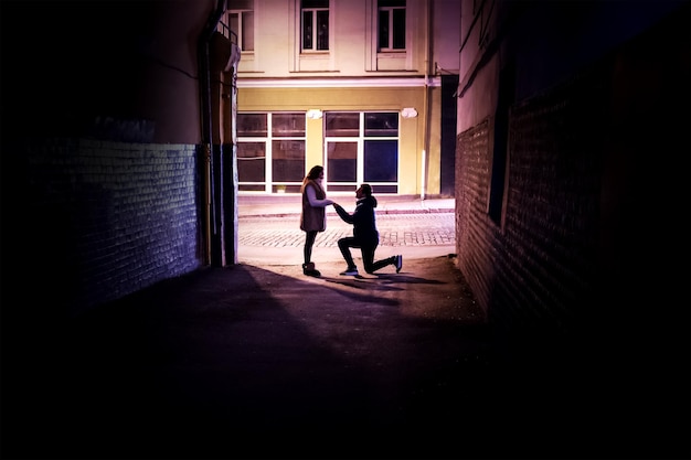 homem faz uma proposta de casamento para uma mulher ajoelhada na luz no final de um beco escuro