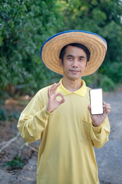 Homem fazendeiro asiático usa camisa amarela, mostra tela do smartphone e dedo bom