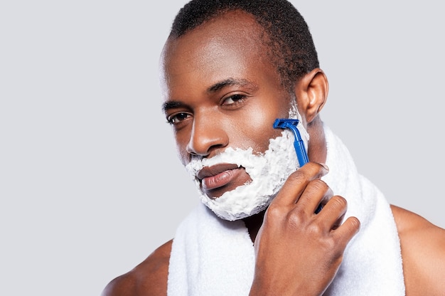 Homem fazendo a barba. Bonito homem africano sem camisa, barbeando o rosto e olhando para a câmera em pé contra um fundo cinza