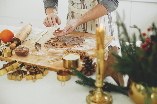 Foto homem fazendo biscoitos de pão de gengibre de natal em close-up em cozinha branca moderna mãos cortando massa de pã de gengibre com cortadores de metal dourados festivos com especiarias e decorações de cozinha