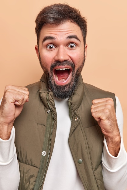 homem fecha os punhos e exclama em voz alta, o gesto de vitória enraíza para o time favorito, enquanto assiste ao jogo, usa camiseta branca e colete expressa emoções positivas