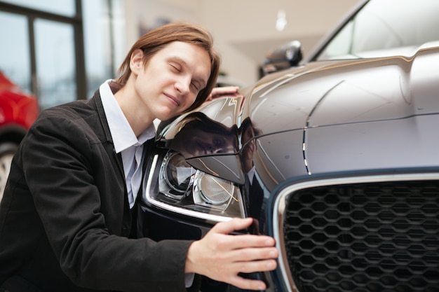 Homem feliz abraçando seu novo automóvel no salão da concessionária, copie o espaço