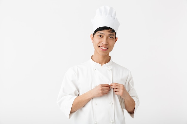 homem feliz asiático com uniforme branco de cozinheiro e chapéu de chef, sorrindo para a câmera em pé, isolado sobre uma parede branca