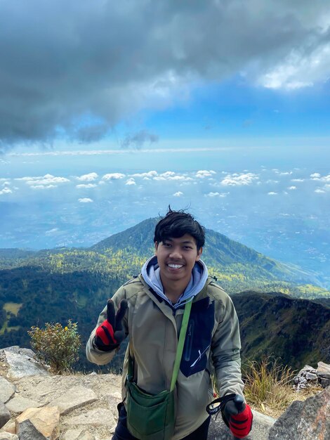 homem feliz asiático de pé na borda do pico da montanha Arjuno com rosto sorridente e fundo de vale