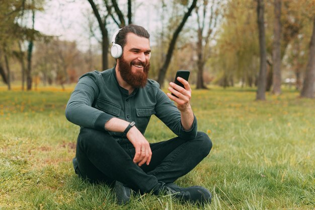 Homem feliz barbudo hippie sentado na grama do parque ouvindo música e usando o smartphone