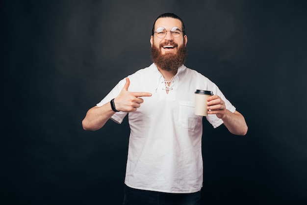 Homem feliz barbudo hippie sorrindo e apontando para a xícara de café de papel para ir