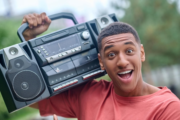 Homem feliz. Cara alegre afro-americano com a boca aberta segurando o toca-discos no ombro com entusiasmo olhando para a câmera ao ar livre