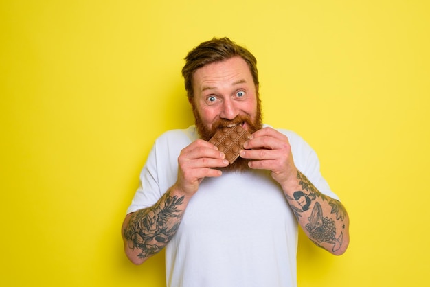 Homem feliz com barba e tatuagens comendo chocolate