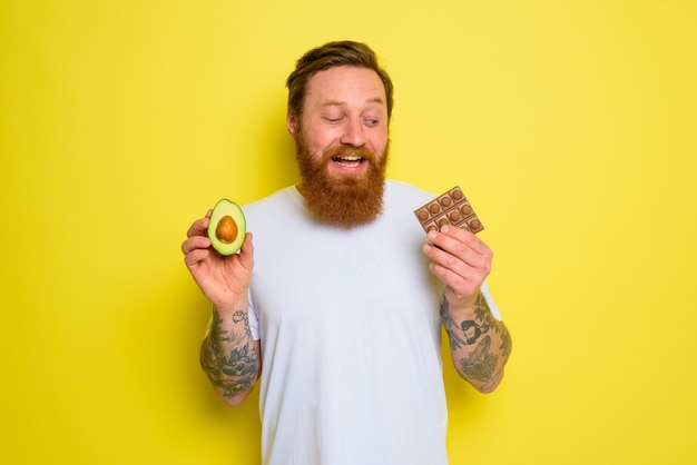 Homem feliz com barba e tatuagens segurando abacate e chocolate