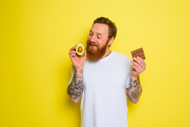 Homem feliz com barba e tatuagens segurando abacate e chocolate