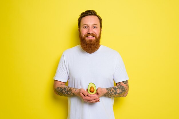 Homem feliz com barba e tatuagens segurando um abacate