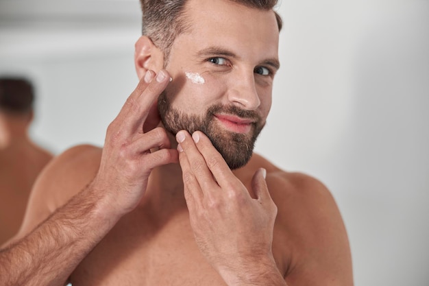 Homem feliz com barba elegante aplica creme antienvelhecimento nos pés de corvo no rosto no banheiro