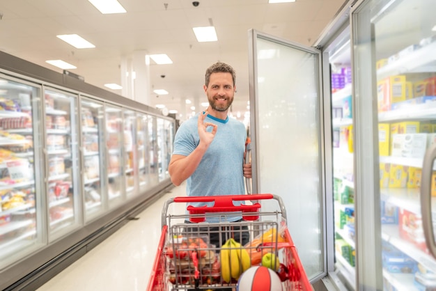 Homem feliz com carrinho de compras comprando comida na mercearia mostrando gesto okey, mercearia