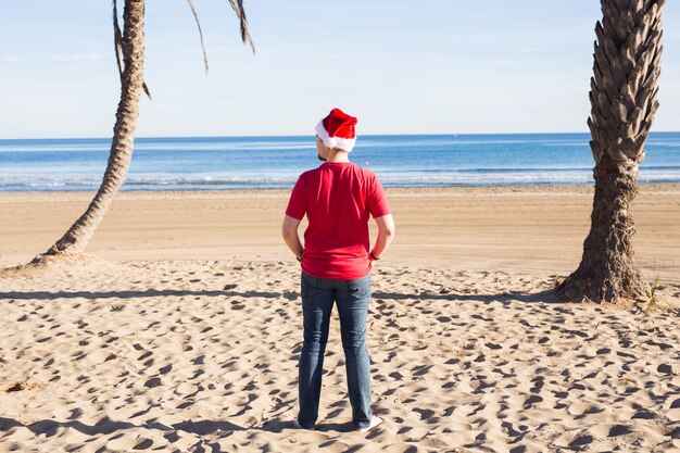 Homem feliz com chapéu de Natal olhando para você enquanto está na praia