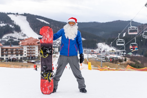 homem feliz com chapéu de Papai Noel vermelho de Natal com snowboard na colina de montanhas de inverno.
