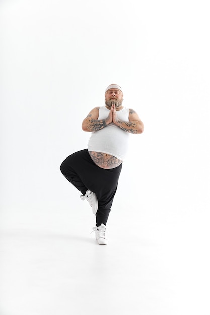 Homem feliz com excesso de peso, barriga grande e tatuagens em roupas esportivas, posando em fundo branco