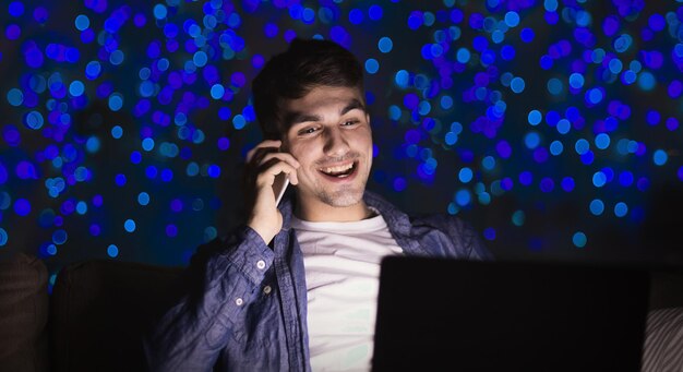 Homem feliz com laptop e smartphone à noite. Rapaz trabalhando até tarde no escritório em casa no bokeh de fundo, copie o espaço