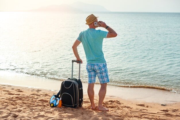 Homem feliz com mala e celular em pé na praia freelancer.