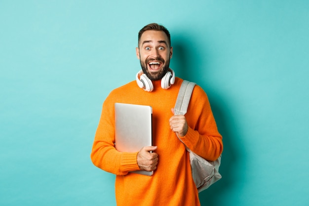 Homem feliz com mochila e fones de ouvido, segurando laptop e sorrindo, parecendo animado, em pé sobre fundo turquesa.