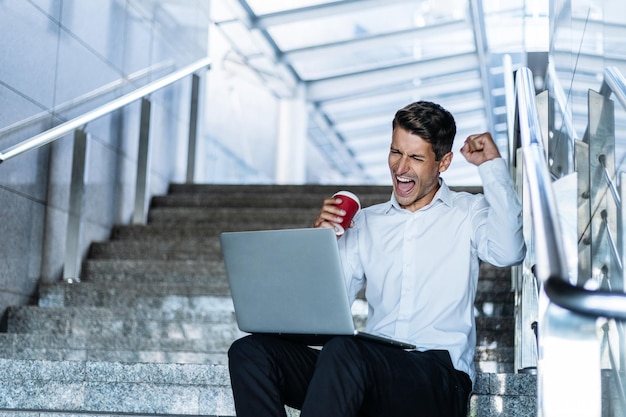 Homem feliz com um café para viagem olhando para a tela do seu laptop