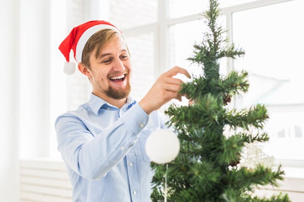 Homem feliz decorando a árvore de Natal em casa com chapéu de Papai Noel. Homem decorando árvore com bugigangas