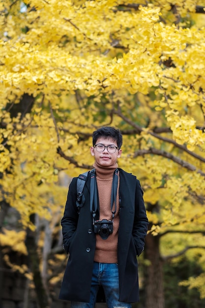 Homem feliz desfruta no parque ao ar livre na temporada de outono viajante asiático de casaco e câmera contra o fundo Yellow Ginkgo Leaves