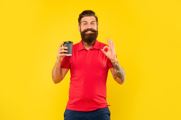 Homem feliz em camiseta vermelha casual dando OK segurando copo descartável fundo amarelo copo de café