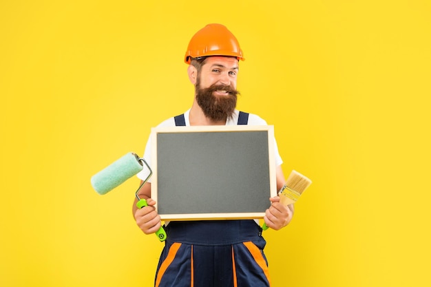 Homem feliz em capacete e roupas de trabalho segura pincel de rolo de pintura e quadro-negro com espaço de cópia