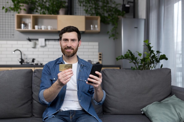 Homem feliz em casa sentado no sofá fazendo compras na loja online usando o aplicativo no telefone e um cartão de crédito bancário