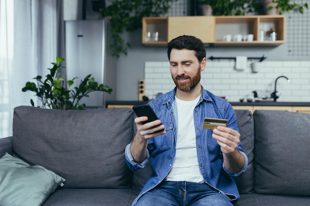 Homem feliz em casa sentado no sofá fazendo compras na loja online usando o aplicativo no telefone e um cartão de crédito bancário