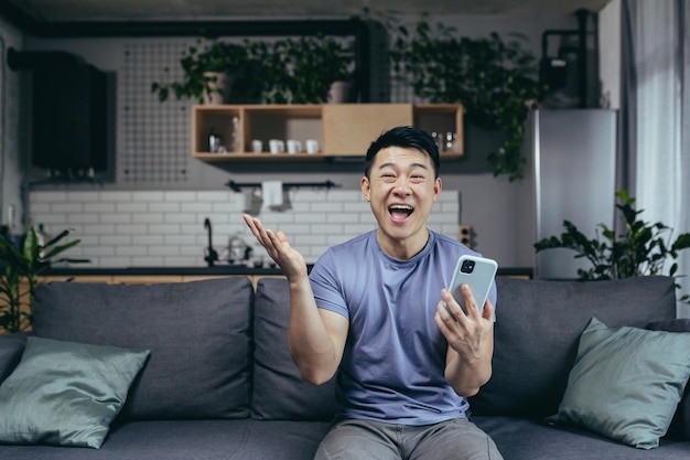 Homem feliz em casa, vitória bem-sucedida, online, sentado no sofá, asiáticos gritam de alegria e sorrisos, olham para a câmera segurando o telefone