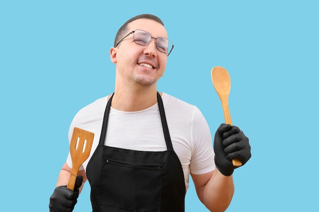 Homem feliz em um avental e colheres de pau em suas mãos em um conceito de culinária de fundo isolado azul