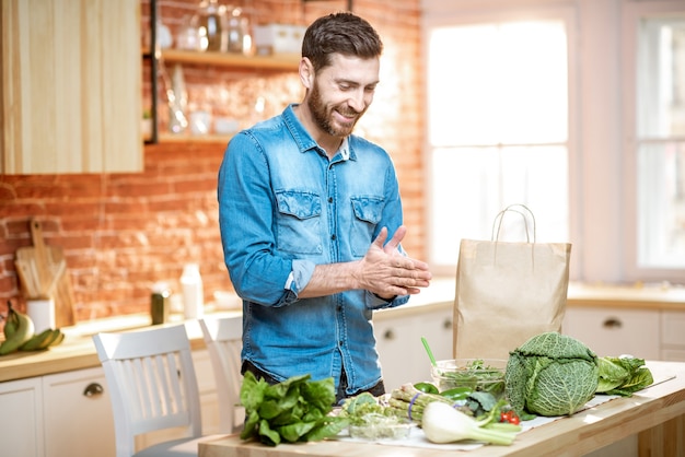 Homem feliz em uma camisa azul com emoções animadas em pé perto da mesa cheia de vegetais verdes frescos na cozinha