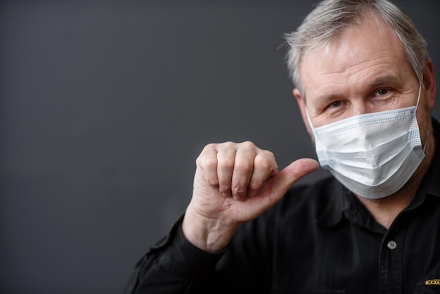 Homem feliz em uma máscara facial estéril para escapar do coronavírus covid19 durante a pandemia de quarentena, mostrando o polegar no topo em um fundo escuro