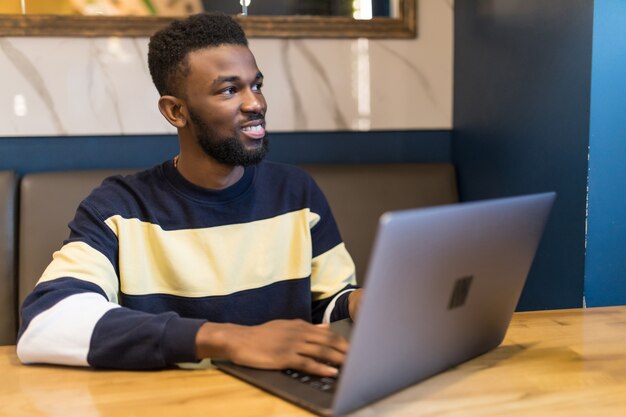 Homem feliz escrevendo um texto no laptop em um café moderno. estudante africano feliz navegando na web no laptop