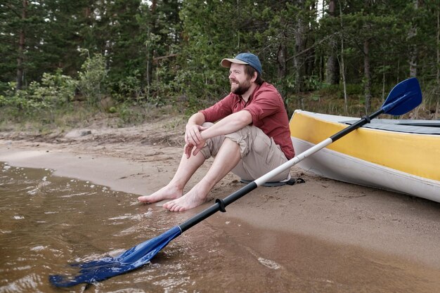 Homem feliz está descansando na margem do lago depois de andar de caiaque, ele olha para longe e sorri