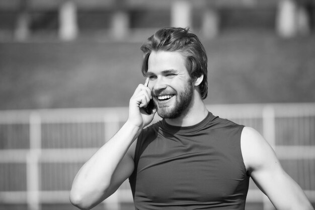 Homem feliz fala no celular na comunicação ensolarada ao ar livre Homem barbudo sorri com smartphone vida moderna Esportista na moda de camiseta esporte azul Moda esportiva e estilo de vida saudável