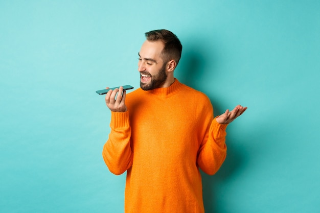 Homem feliz falando no viva-voz, gesticulando e gravando mensagem de voz no celular, em pé com a camisola laranja sobre fundo azul claro.