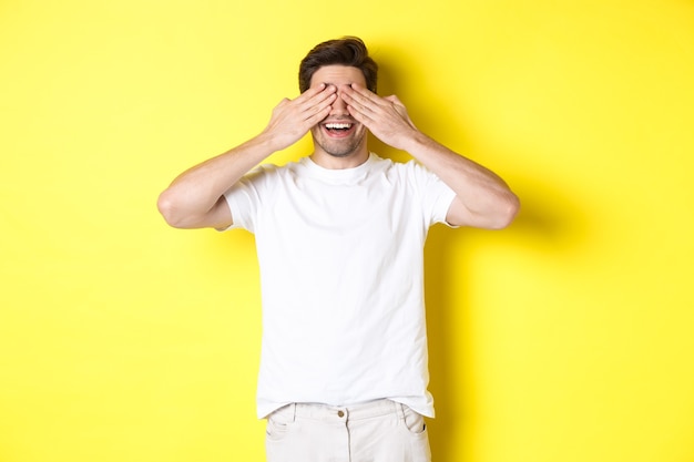 Homem feliz fechando os olhos e esperando a surpresa, sorrindo divertido, em pé sobre a parede amarela