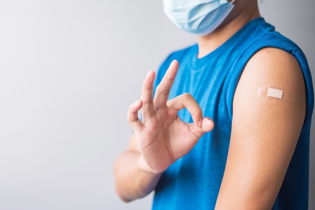 Homem feliz, mostrando o braço com curativo e dando sinal de mão ok após receber a vacina. Vacinação, imunização, inoculação e pandemia de Coronavírus (Covid-19)