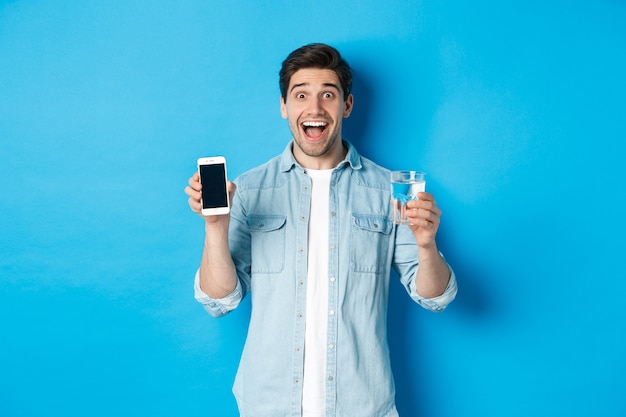 Homem feliz mostrando um copo d'água e a tela do celular, recomendando o aplicativo de saúde para smartphone, em pé sobre um fundo azul