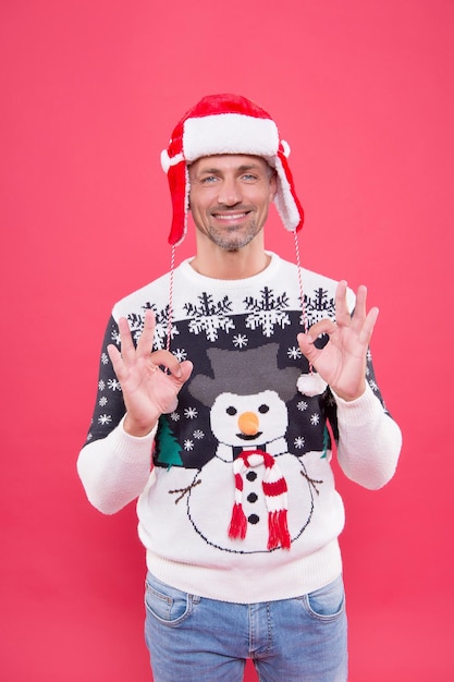 Homem feliz mostrar o gesto certo, vestindo o suéter de malha engraçado e um chapéu para comemorar as férias de Natal.