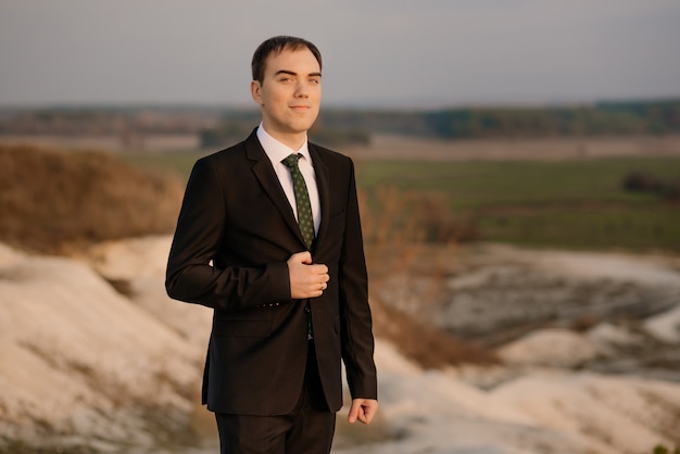 Foto homem feliz, noivo de terno elegante casaco ao ar livre em fundo natural. noivo elegante esperando a noiva. retrato emocional do noivo ao ar livre ao pôr do sol