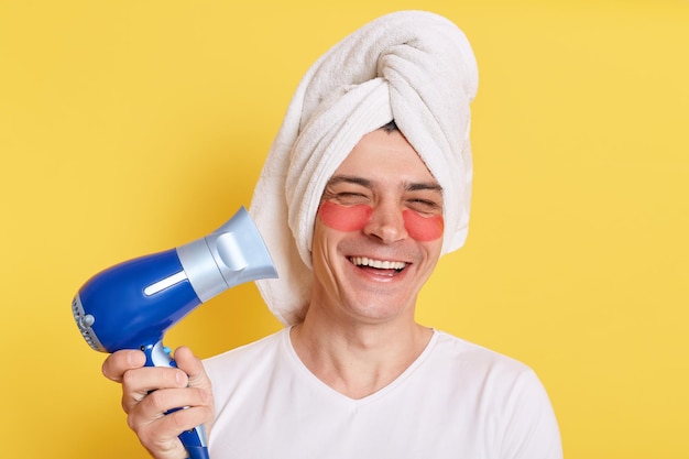 Foto homem feliz otimista enrolado toalha na cabeça fazendo procedimentos de cosmetologia matinal secando o cabelo com secador de cabelo rindo alegremente posando com manchas sob os olhos isolados sobre fundo amarelo