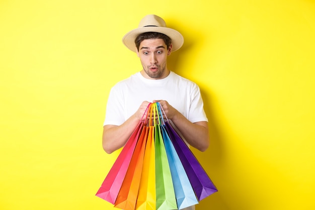 Homem feliz parecendo surpreso com sacolas de compras, comprando lembranças nas férias, em pé sobre a parede amarela