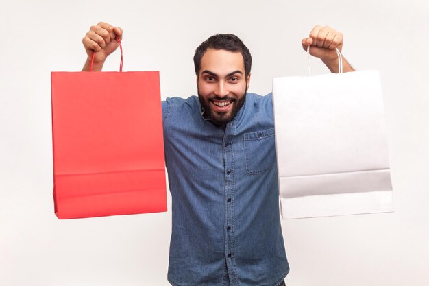 Foto homem feliz positivo na camisa azul mostrando dois saco de papel olhando para a câmera com sorriso, compras bem sucedidas. tiro de estúdio interior isolado no fundo branco